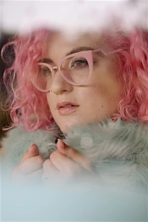 Close-up portrait of a woman with bright pink curly hair wearing pink geometric eyeglasses and a fluffy pale green jacket
