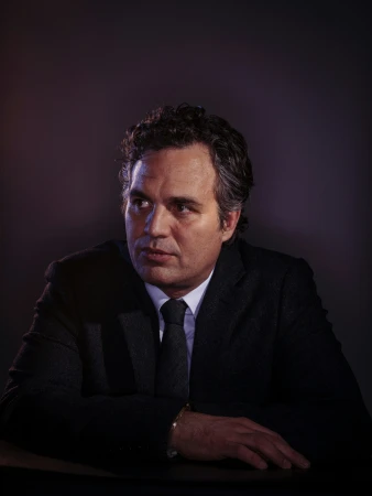 Portrait of Mark Ruffalo wearing a dark suit and tie against a dark background, photographed by Brinson Banks.