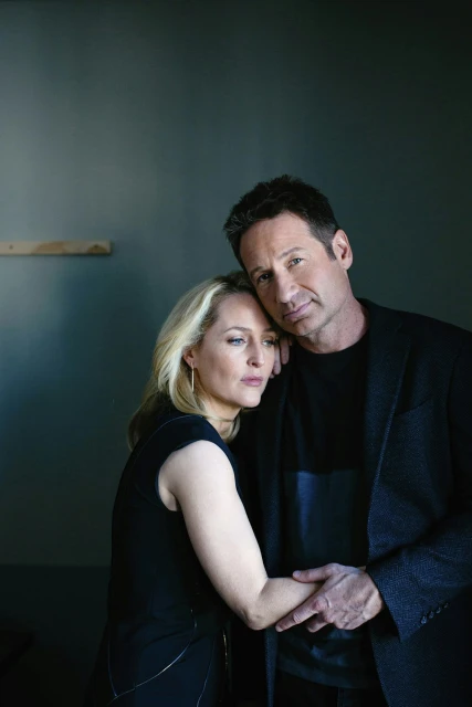 Portrait of actors Gillian Anderson and David Duchovny in dark formal wear, close together indoors against a muted green wall.