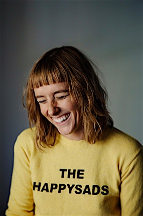 Smiling woman with short hair wearing a yellow sweater with the words 'THE HAPPYSADS' printed on it in a photo studio setting.