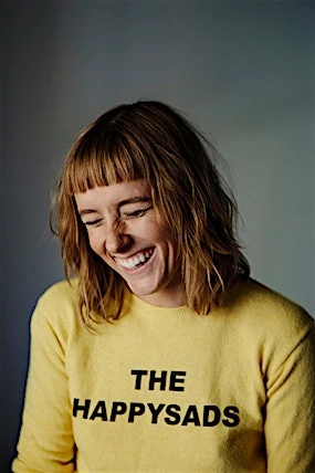 Smiling woman with short hair wearing a yellow sweater with the words 'THE HAPPYSADS' printed on it in a photo studio setting.
