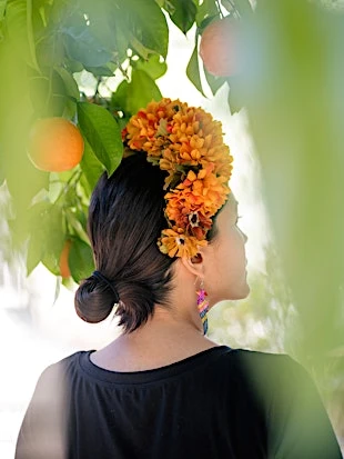 A woman with orange flower crown stands beneath an orange tree in an outdoor portrait, showcasing natural beauty and botanical elements.