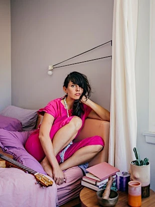 Woman with long hair and bangs in a pink jumpsuit sitting on bed with a ukulele and table with books, brass bowl, and candleholders.