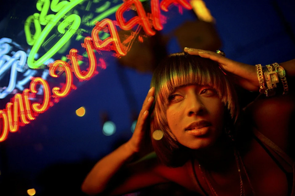 Close-up shot of an adult woman with gold jewelry and neon lights in the background, styled in dark clothing and dramatic lighting.