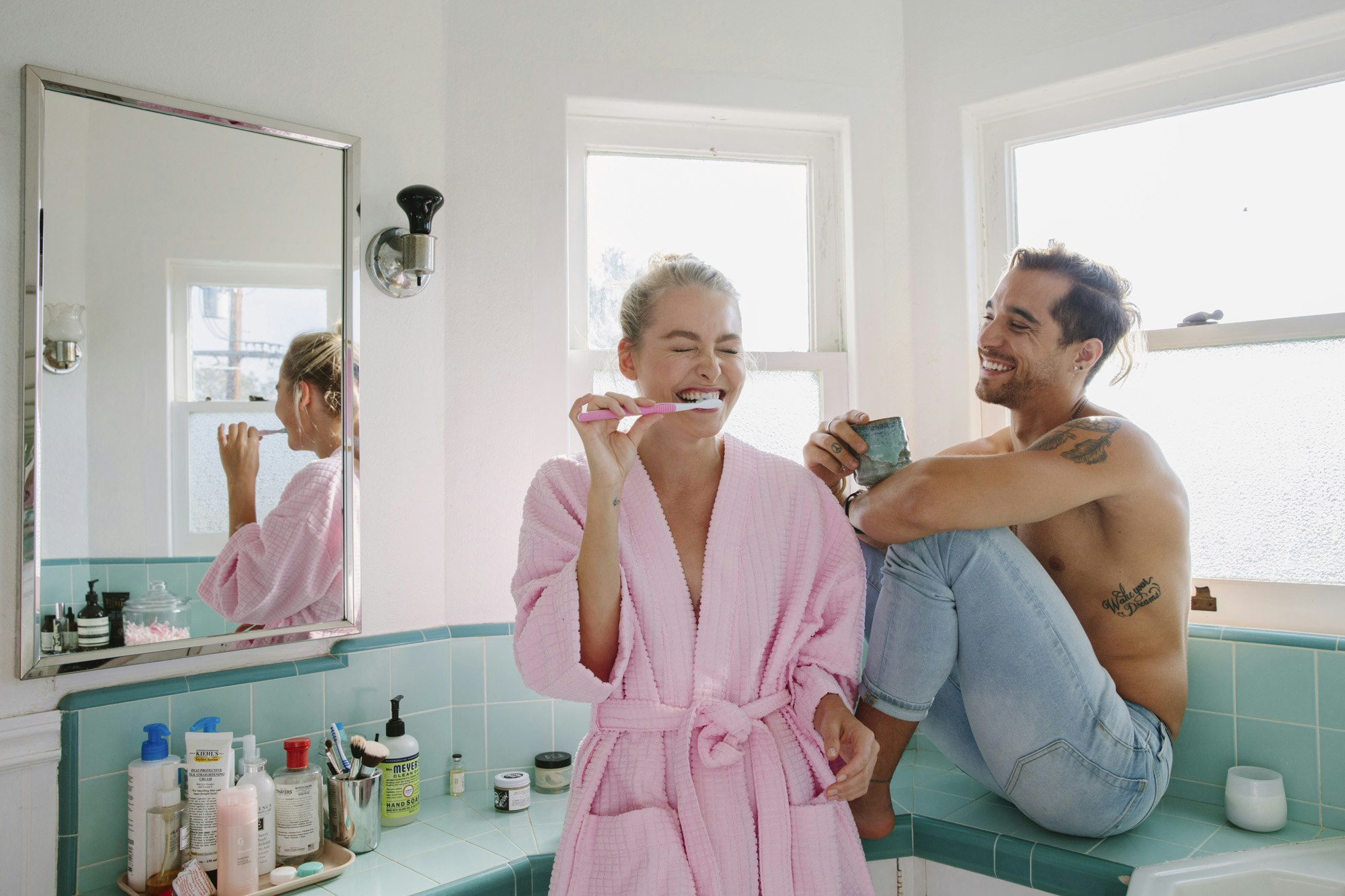 A smiling woman in a pink robe brushing her teeth and a shirtless man in jeans looking at her inside a brightly lit tiled bathroom