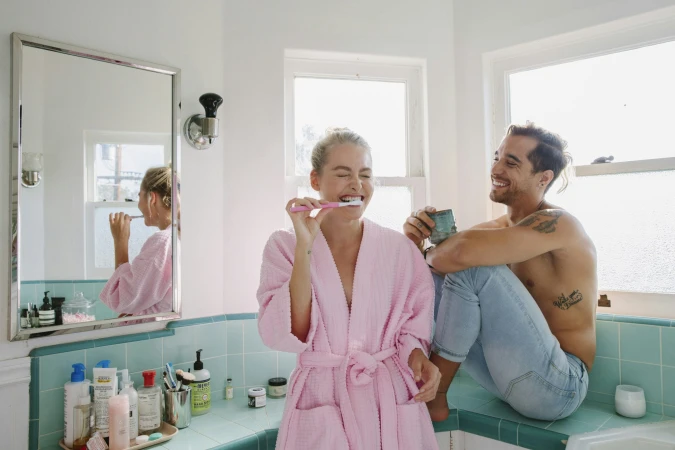 A smiling woman in a pink robe brushing her teeth and a shirtless man in jeans looking at her inside a brightly lit tiled bathroom