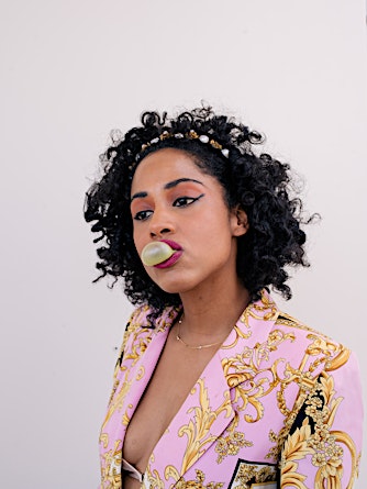Portrait of a woman blowing a bubble with bubble gum, wearing a printed pink blazer and a beaded headband against a white backdrop.