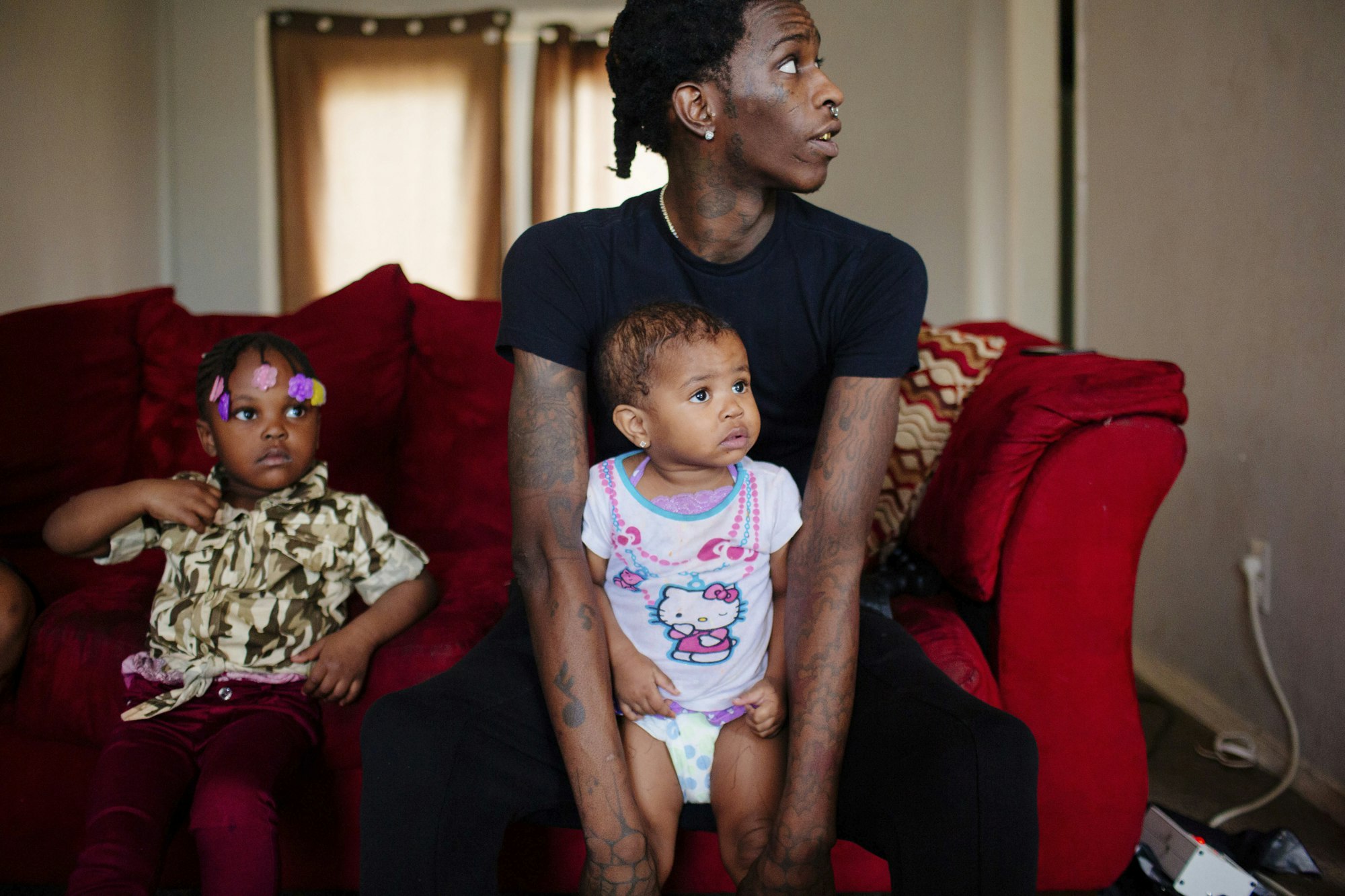 An adult man sits with two young children on a red sofa, with a window in the background, in a domestic interior.