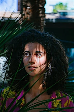 Portrait of a young woman with curly dark hair, wearing a colorful shirt, framed by green plants and looking slightly upwards outdoors.