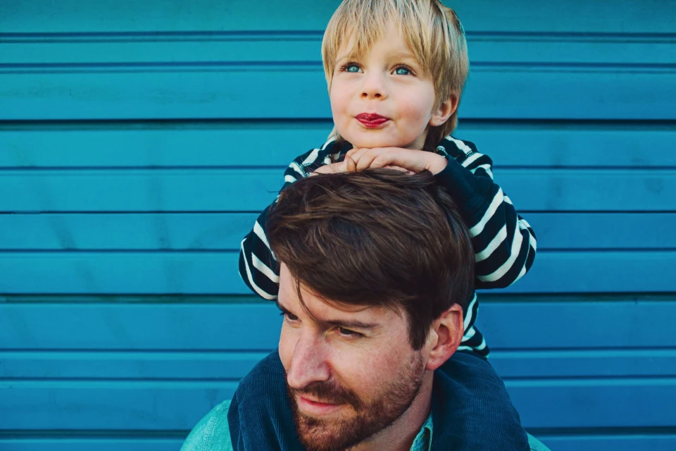 A smiling little boy with blonde hair sits on his father's shoulders, both looking towards the left, against a bright blue wood-paneled background.