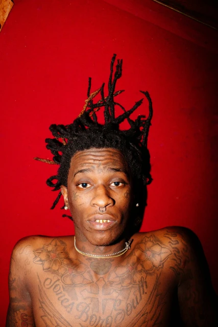 Close-up portrait of a Black adult male with dreadlocks, facial piercings, gold chain, and tattoos set against a vibrant red backdrop.