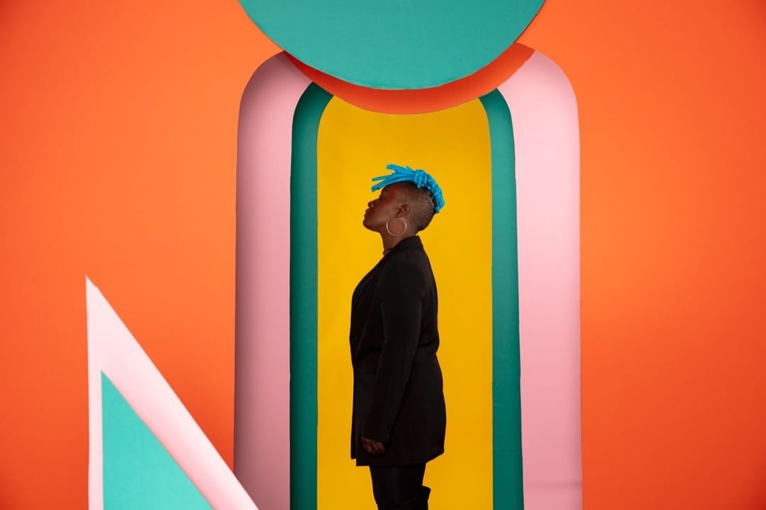 Stylish portrait of a black woman with blue braided hair posing in front of colourful geometric shapes in a bright indoor space.