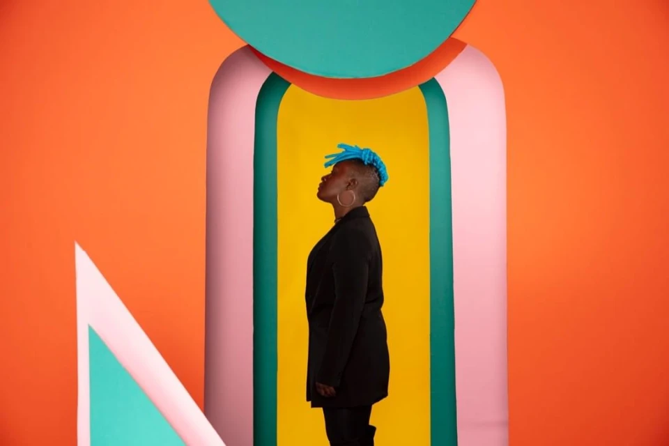 Stylish portrait of a black woman with blue braided hair posing in front of colourful geometric shapes in a bright indoor space.