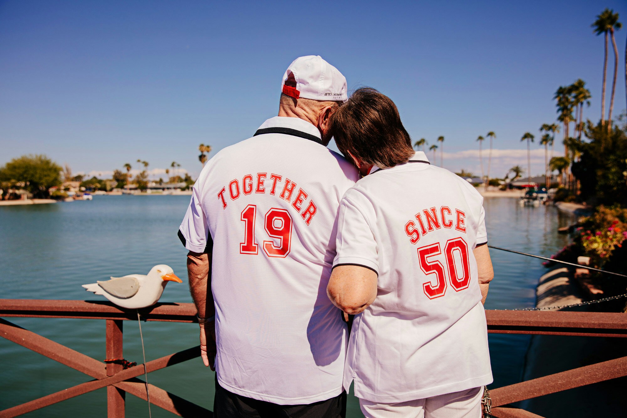 Elderly couple wearing matching jerseys with backs facing camera, standing on a bridge overlooking a lake, with palm trees in background.