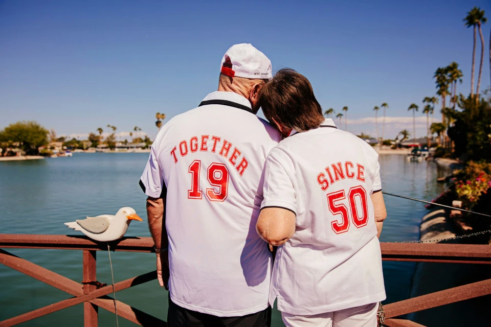 Elderly couple wearing matching jerseys with backs facing camera, standing on a bridge overlooking a lake, with palm trees in background.