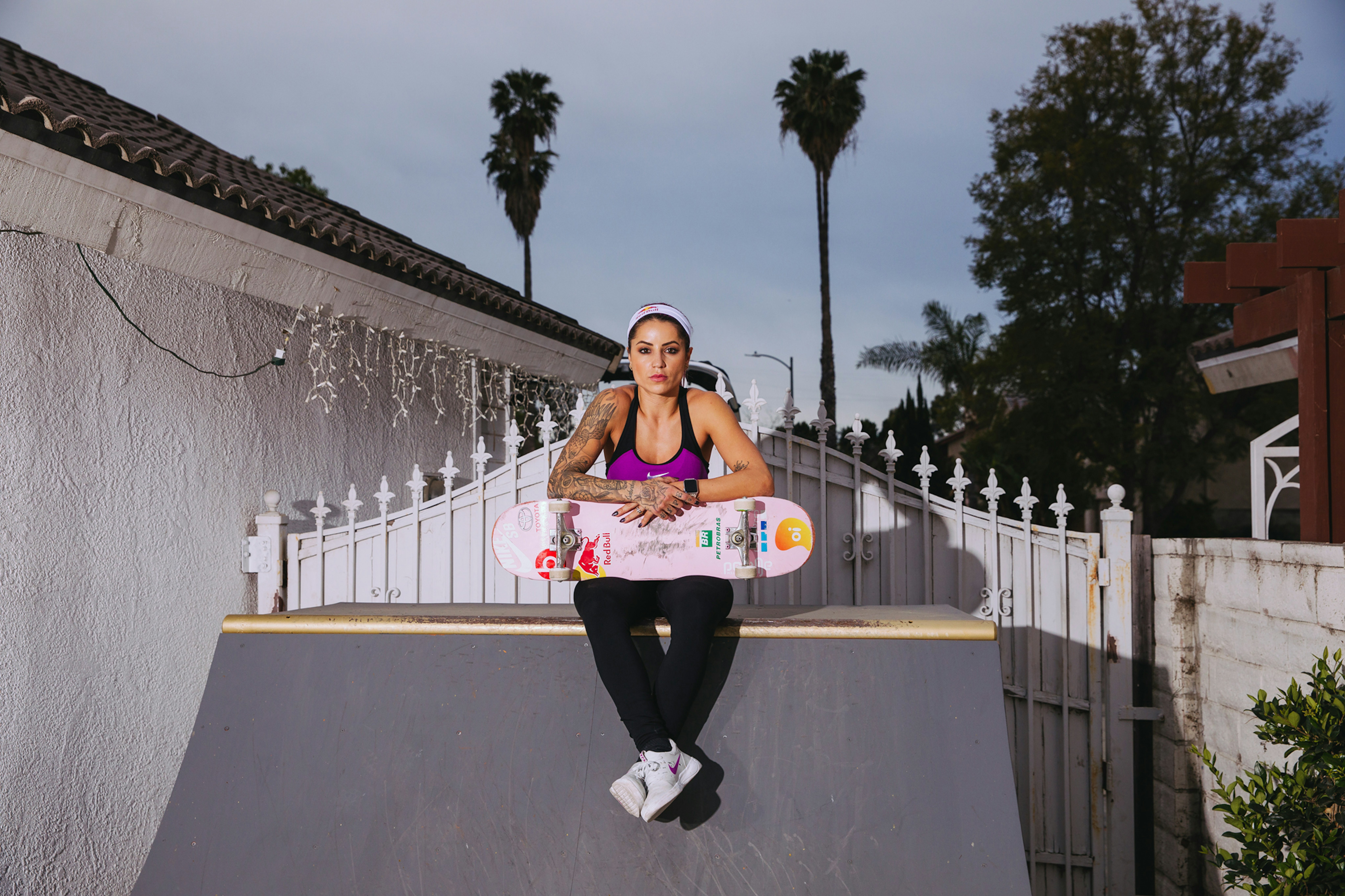 Tattooed woman in athletic wear sits with skateboard on ramp, set against a backdrop of palm trees and residential houses.