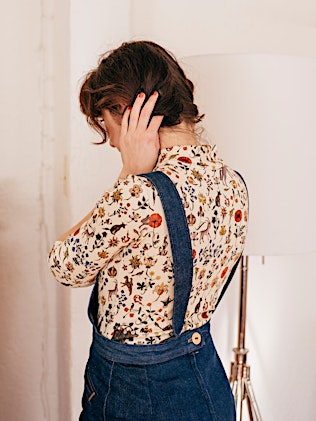 Brunette woman in floral blouse and denim overalls stands indoors with hand to head, in soft, diffused lighting from floor lamp.