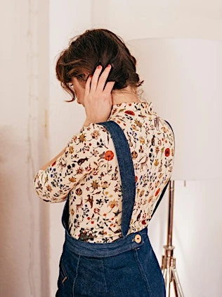 Brunette woman in floral blouse and denim overalls stands indoors with hand to head, in soft, diffused lighting from floor lamp.