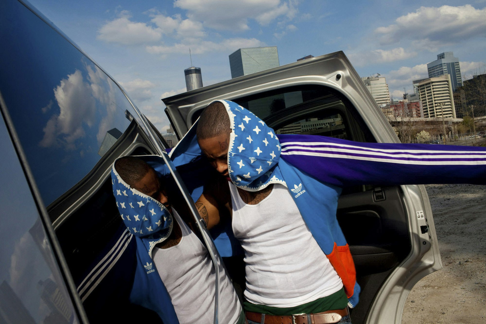 An adult male in a blue and white track jacket stands next to the open door of a silver SUV, with city buildings in the background under a blue sky.