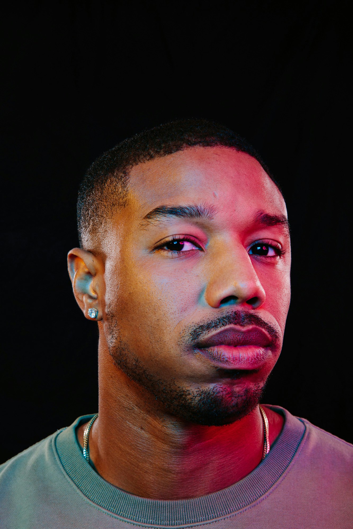 Headshot of an adult male of African descent with short hair, wearing a grey shirt and gold chain, set against a black background.