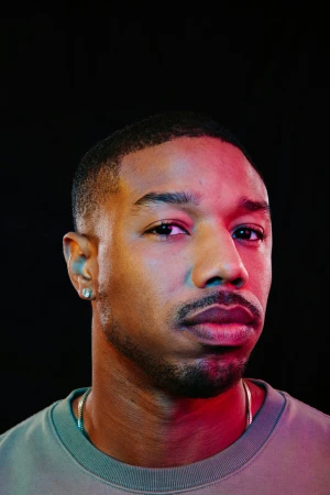 Headshot of an adult male of African descent with short hair, wearing a grey shirt and gold chain, set against a black background.
