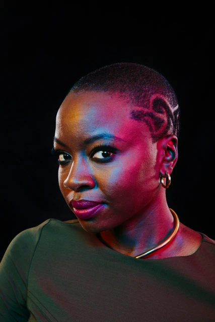 Close-up portrait of a Black woman with short hair, custom design, gold necklace, wearing a green top, against a dark background.