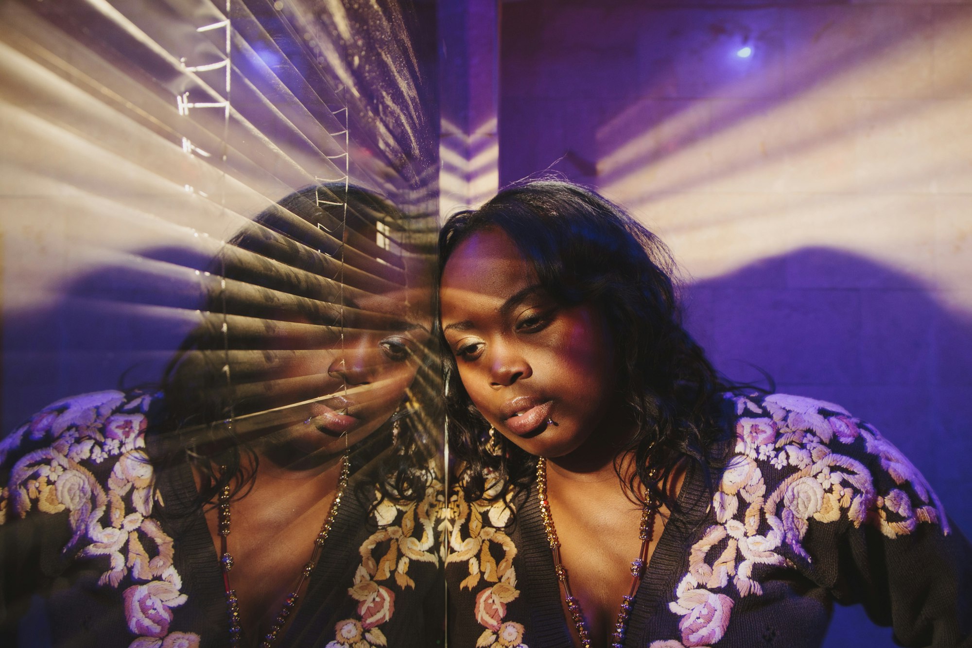 Portrait of a woman with a mirrored reflection, illuminated by blue and purple lighting, wearing a floral-patterned top and beaded necklace.