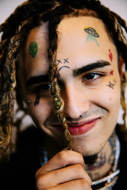 Close-up portrait of a smiling adult male with dreadlocks, face tattoos, and multiple piercings, set against a neutral background.