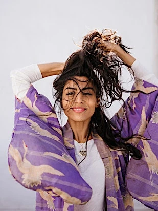 Smiling woman with dark hair holds her hair up, wearing a printed purple kimono, posing against a white background.