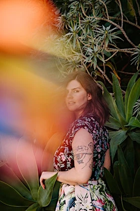 Woman with floral top and skirt poses surrounded by green plants with a light leak effect casting rainbow colors on the left side.