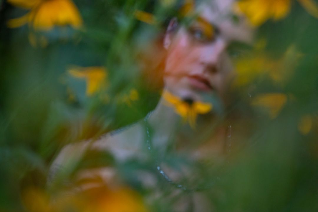 Abstract portrait of a young adult peering through yellow flowers and green foliage in a blurred, outdoor setting with soft lighting.
