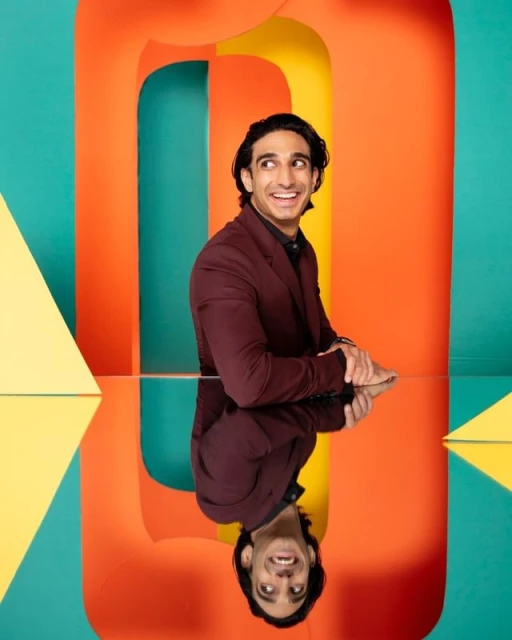 Person in a suit reflected on a glass table, with colorful geometric background shapes in orange, green, and yellow.