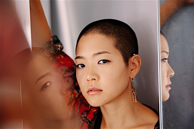 Close-up portrait of a woman with a buzzcut and earrings looking directly at the camera between reflective mirrors.