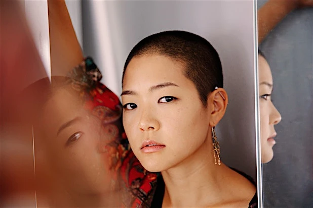 Close-up portrait of a woman with a buzzcut and earrings looking directly at the camera between reflective mirrors.