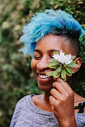 Smiling woman with blue hair, nose ring, and flower in hand poses outdoors against a green leafy background in a medium shot.