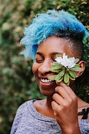Smiling woman with blue hair, nose ring, and flower in hand poses outdoors against a green leafy background in a medium shot.