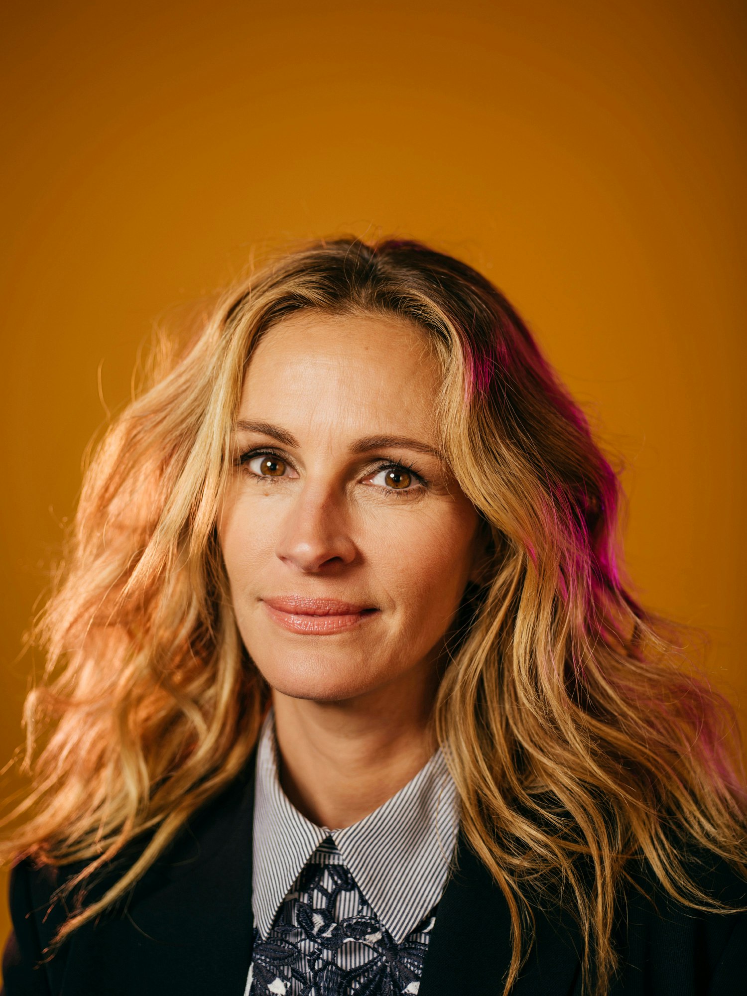 Portrait of Julia Roberts with long blonde hair wearing a dark blazer and patterned shirt against a mustard yellow background
