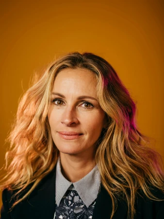 Portrait of Julia Roberts with long blonde hair wearing a dark blazer and patterned shirt against a mustard yellow background