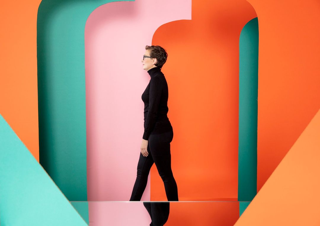 Side view of a woman in a black outfit walking against colorful orange, pink, and teal geometric shapes in an indoor setting.