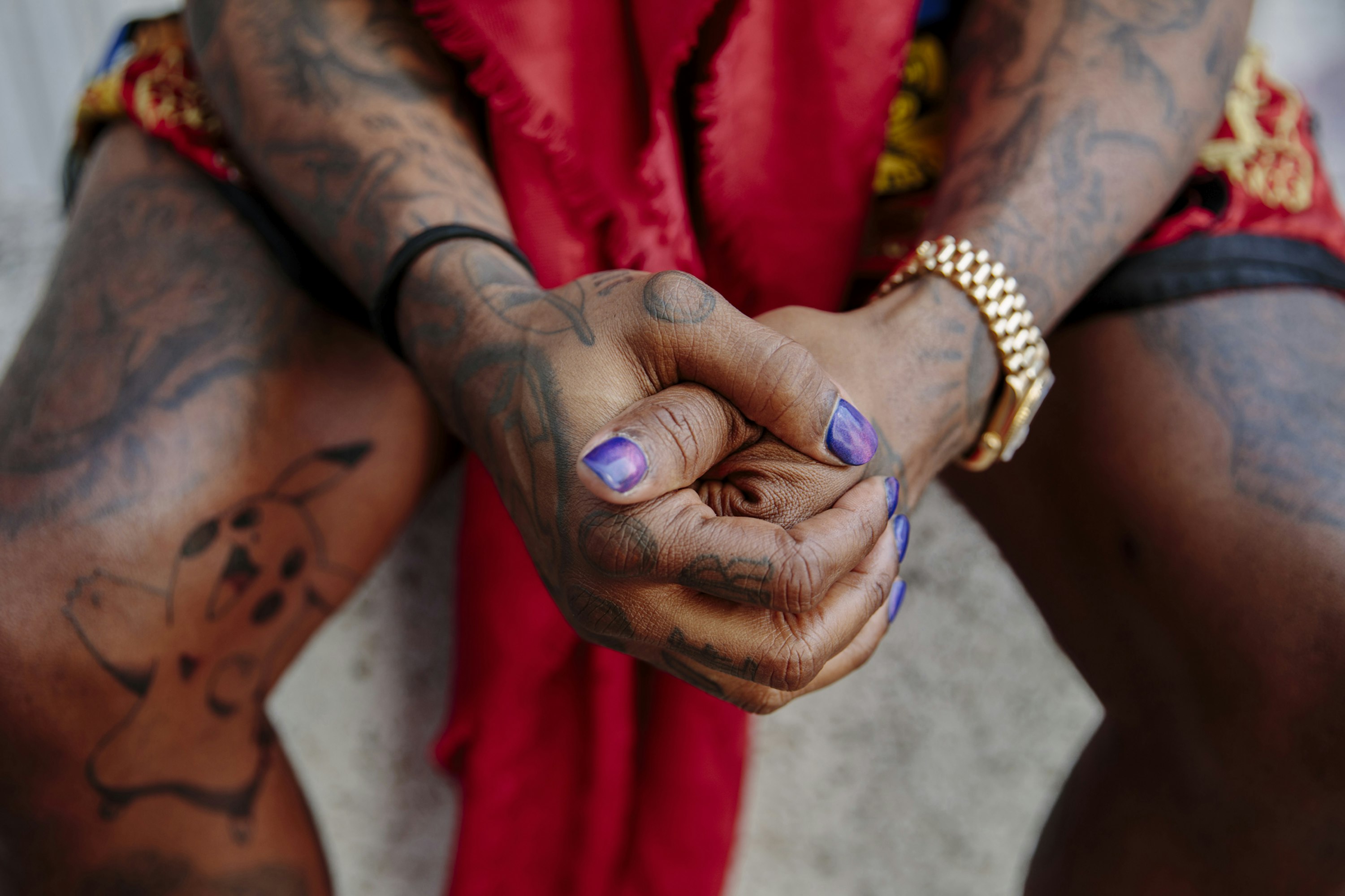 Close-up of a person with heavily tattooed arms and legs, manicured fingernails, gold watch, seated with hands clasped on laps, outdoors.