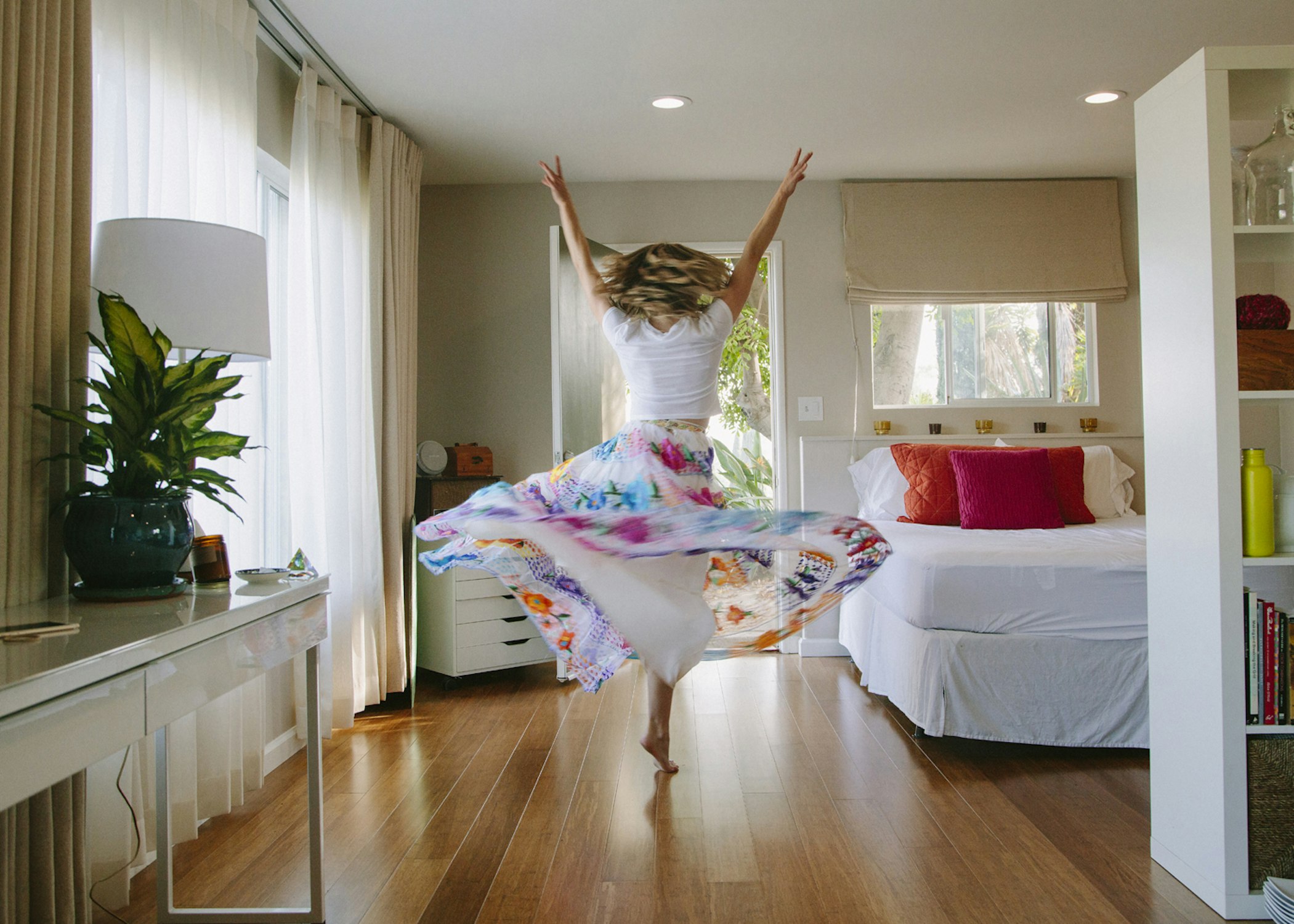 Woman in colorful skirt dancing barefoot in a bedroom with a bed, desk, window and curtains, bathed in soft, natural light.