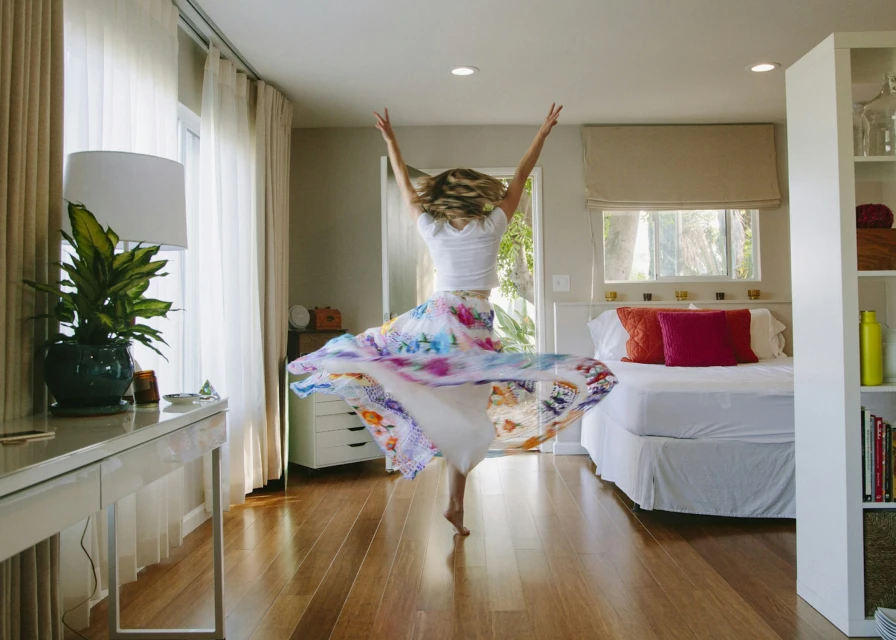 Woman in colorful skirt dancing barefoot in a bedroom with a bed, desk, window and curtains, bathed in soft, natural light.