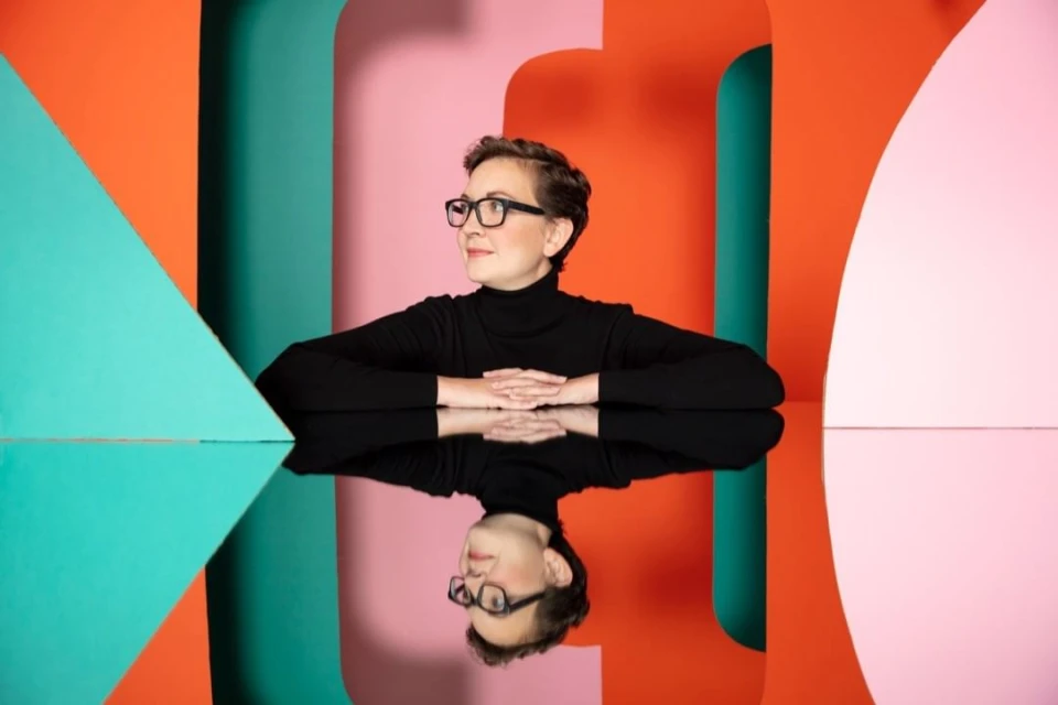 Woman in black turtleneck and glasses sitting at table with reflection, colorful geometric background. Studio portrait, looking away.