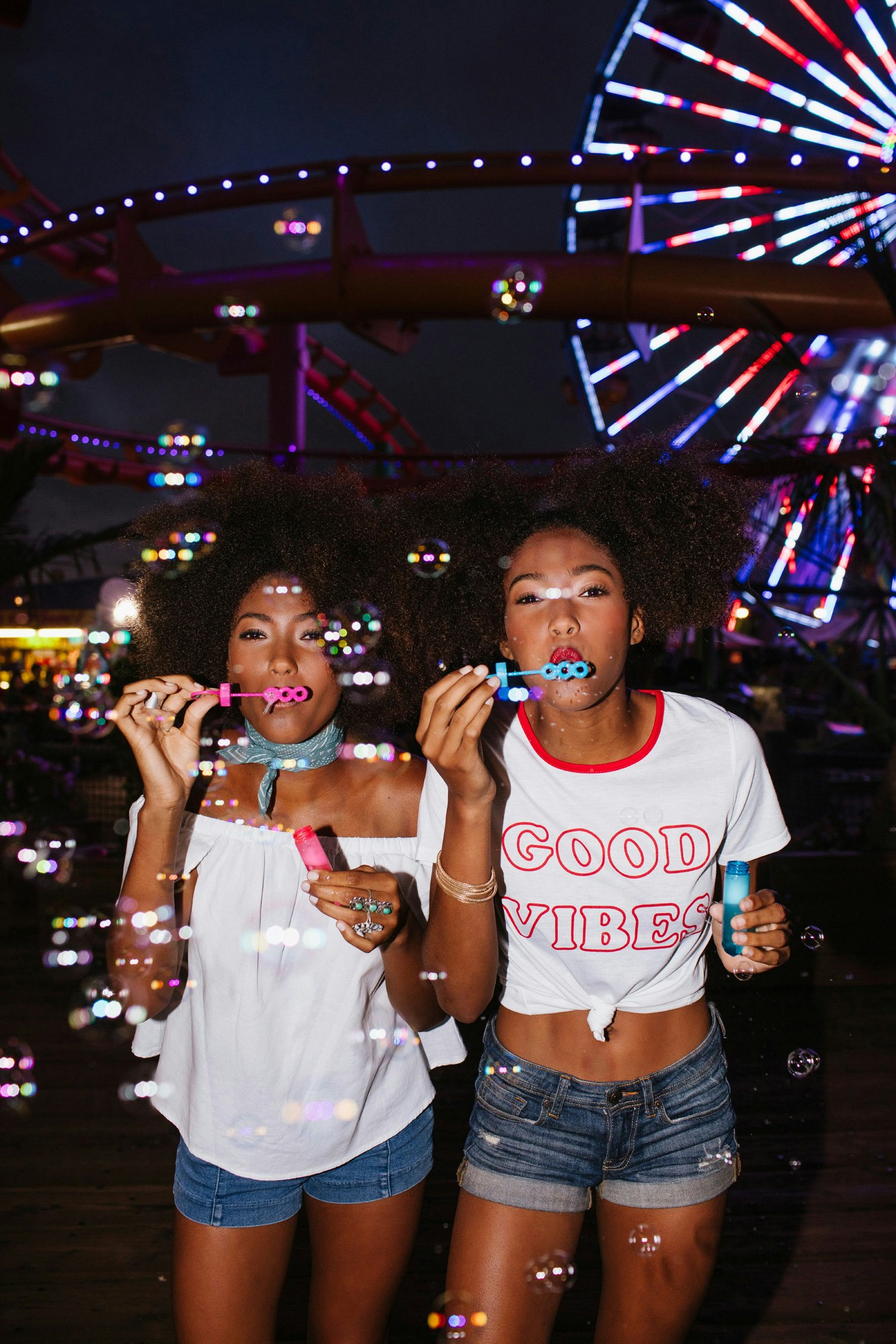 Two young African-American women blowing bubbles at night in front of a brightly lit Ferris wheel, wearing casual summer clothes and jewelry.