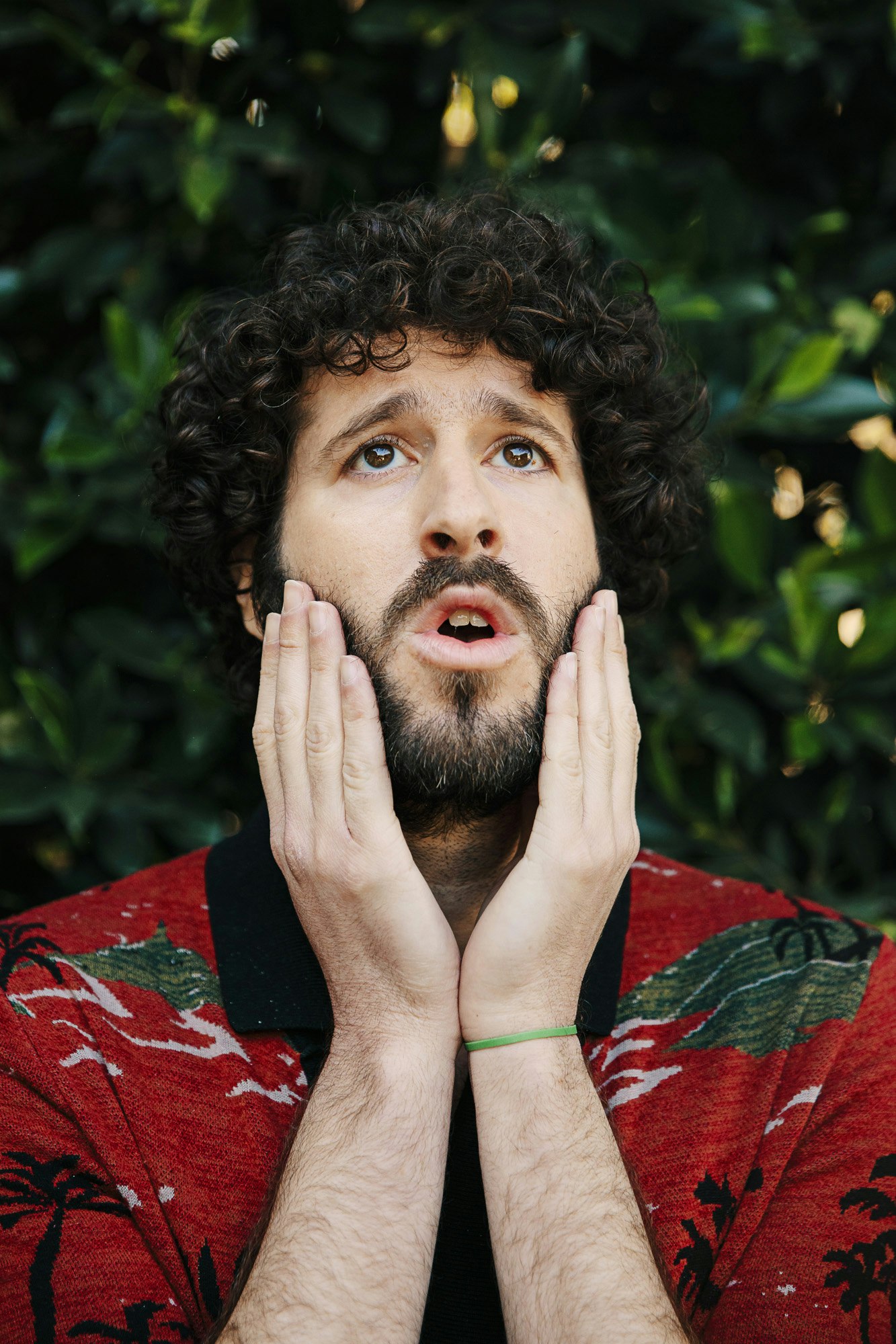 Surprised man with curly hair and beard looks upward, palms on face, wearing a red palm-tree patterned shirt against a green foliage background.
