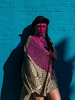 Woman in patterned kimono poses against a bright turquoise brick wall with a shadow cast on the side of her face and the wall.