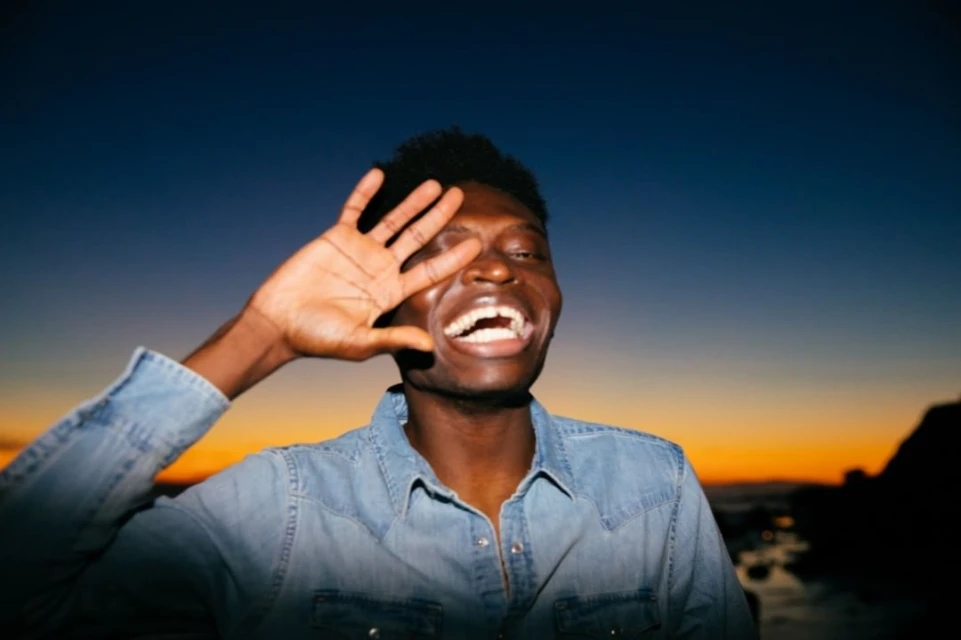 A close-up shot of a smiling young black man in a denim shirt, shielding his eyes with his hand against a vibrant sunset sky backdrop.
