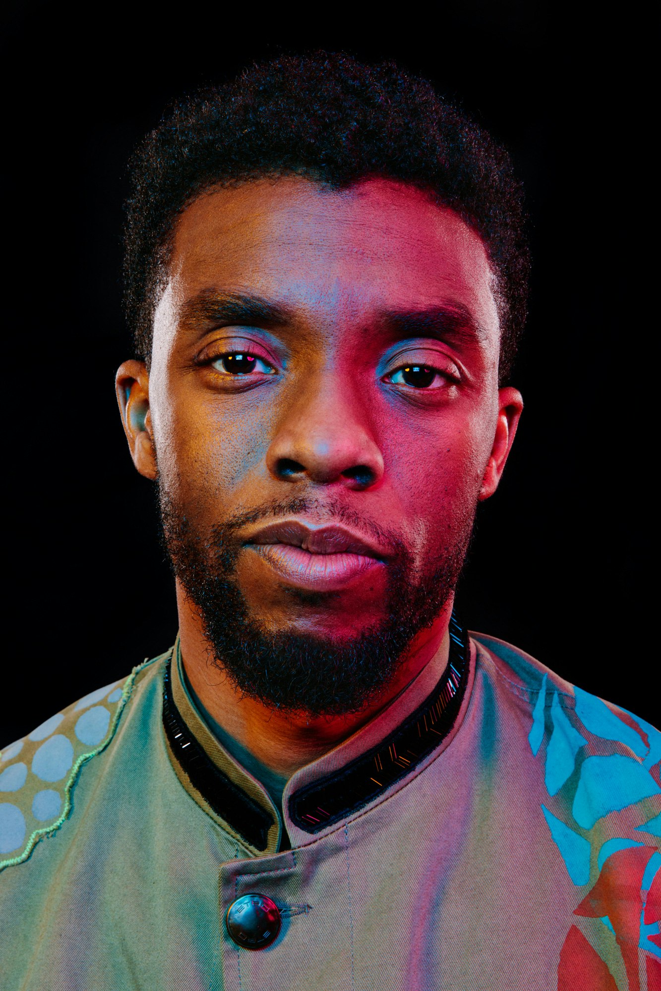 Portrait of Chadwick Boseman with dark hair and serious expression, lit with blue and red lighting against a dark backdrop.