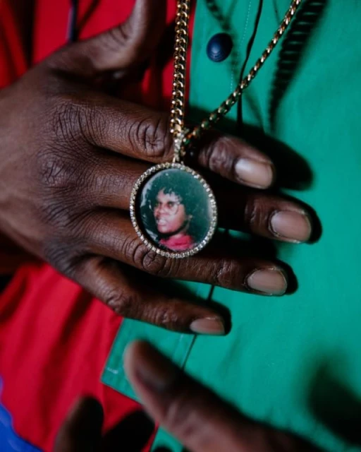 Person wearing a gold chain with a round medallion featuring a portrait, held against a green and red shirt.