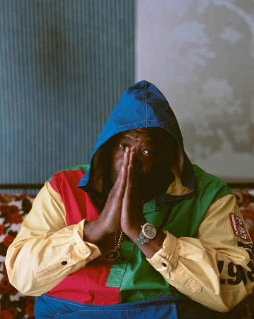 Portrait of an adult male wearing a colorful hooded jacket, with hands clasped in front of him and sitting indoors against a striped wall.